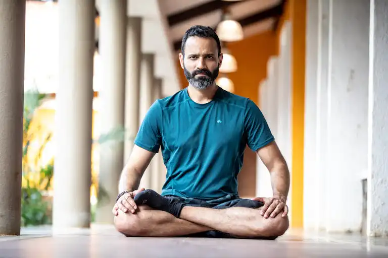 A man with a beard sitting in a yoga pose indoors, exuding calm and focus.